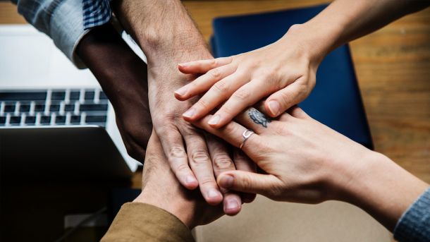 Mehrere Menschen legen ihre Hände übereinander als Zeichen von Teamwork und Zusammenhalt, im Hintergrund ein Laptop und ein Holztisch.